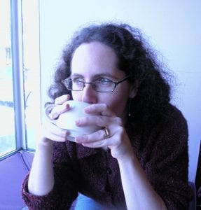 Miriam Axel-Lute, the editor of Shelterforce magazine, drinks from a white mug as she stares out to the left. She is wearing glasses and sitting next to a window.