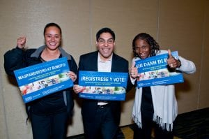 Three members of the Non-Profit Housing Association of Northern California give a thumbs up as they hold "Register and Vote" signs in different languages.