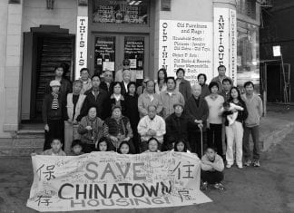 A black and white photo of a dozen or so residents of a multifamily building standing outside with a "Save Chinatown Housing" sign.