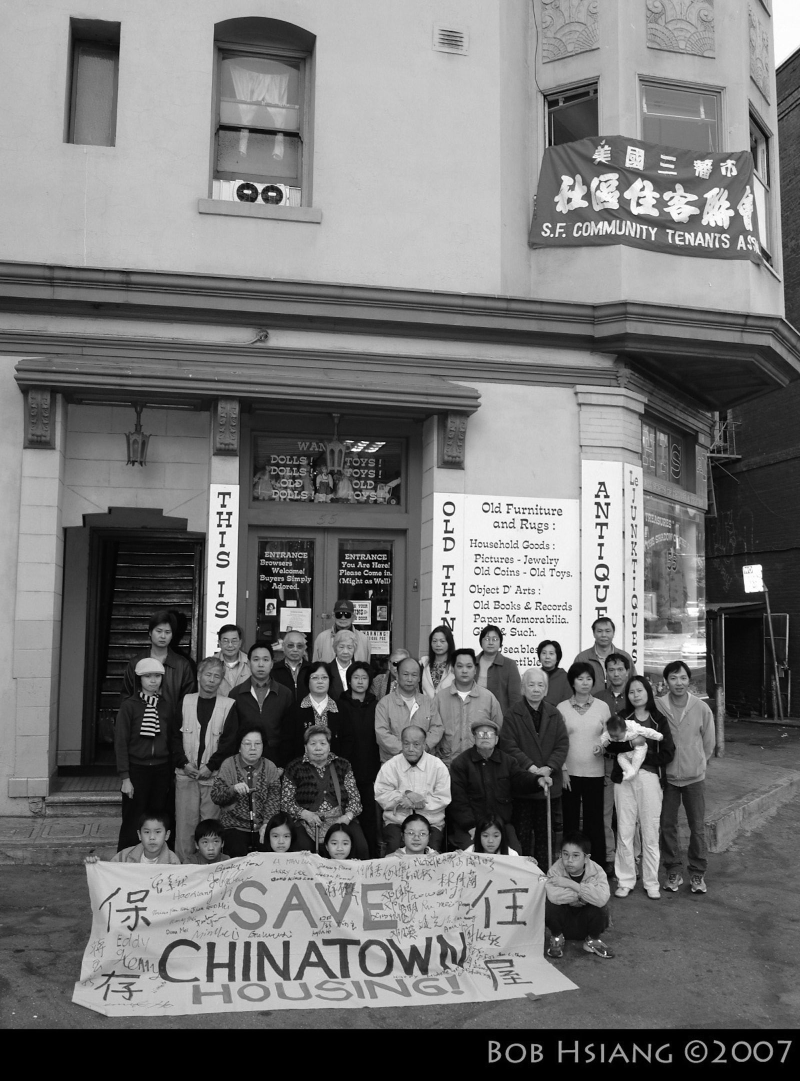 A black and white photo of a dozen or so residents of a multifamily building standing outside with a "Save Chinatown Housing" sign.