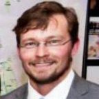 Jarrett Murphy, executive editor and publisher of City Limits, smiles at the camera. He is wearing a pair of glasses, a white top and a grey colored jacket.