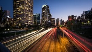 Taken during the evening, this image is of Downtown Los Angeles and shows buildings with lights. While you cannot see cars on a highway, you can see the white and red lines of lights, which shows movement. Illustrating an article about the 30 percent AMI threshold