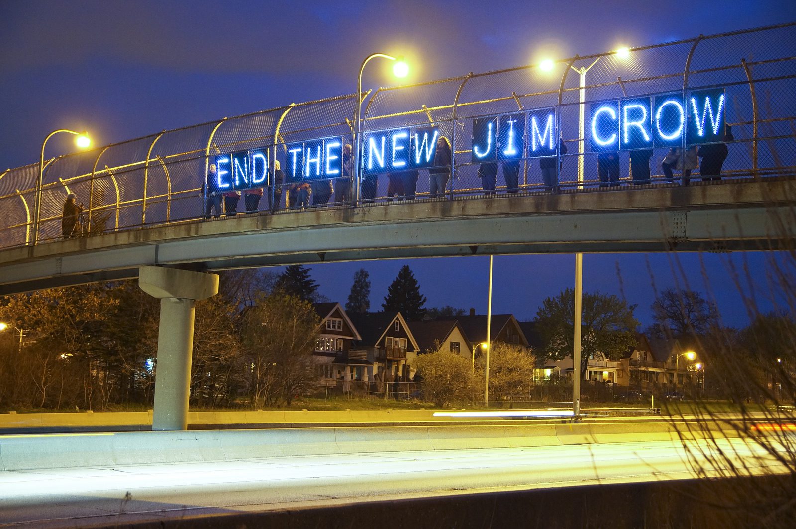 On an overpass over a highway, people hold a lighted sign that reads "End the New Jim Crow."