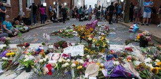 A street is covered with flowers and photos of Heather Heyer, as people look on the background