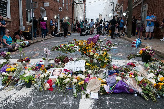 A street in Charlottesville is covered with flowers and photos of Heather Heyer as people look on the background. The word "peace" is chalked on the pavement in the foreground and a sign reads "No Place for Hate."
