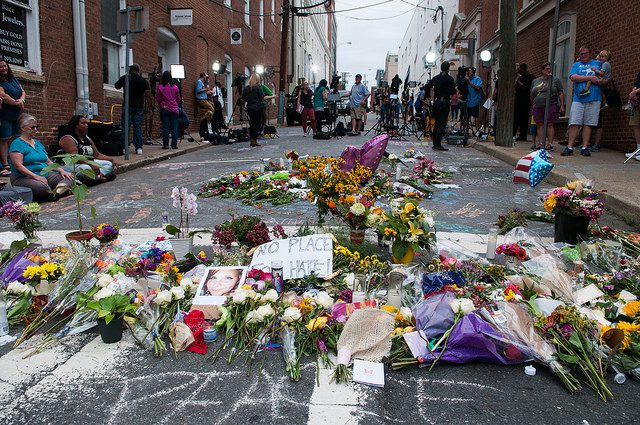 A street is covered with flowers and photos of Heather Heyer, as people look on the background