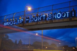 Neon "Hands Up-Don't Shoot" sign displayed on highway overpass.