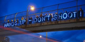 Neon "Hands Up-Don't Shoot" sign displayed on highway overpass.