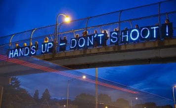 Neon "Hands Up-Don't Shoot" sign displayed on highway overpass.