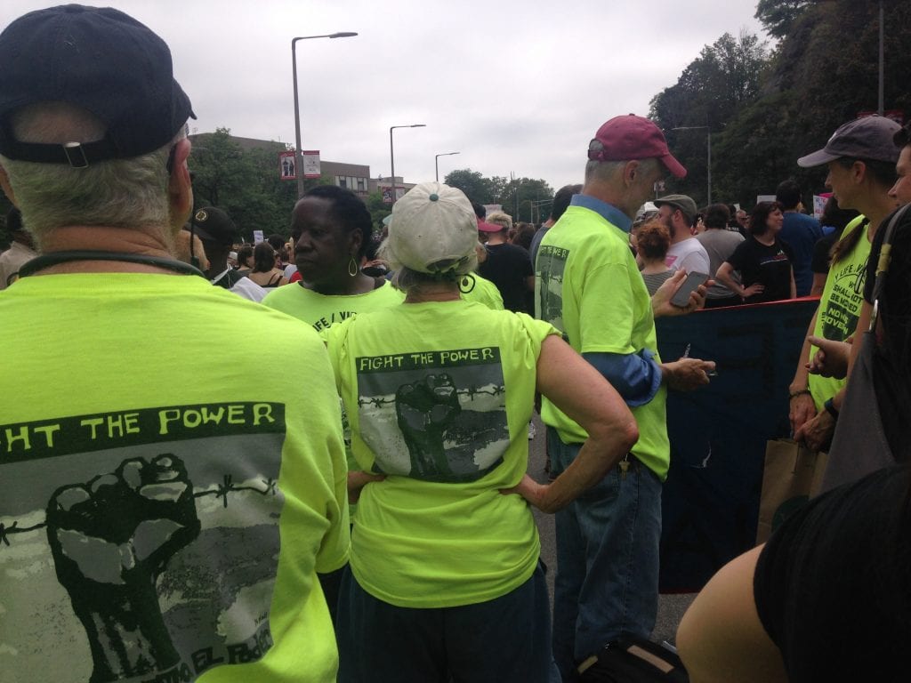 Several people seen from behind in a crowd wearing neon yellow shirts that read "Fight the power" over a picture of a raised fist closed around barbed wire.