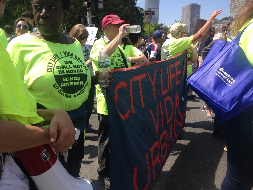 A dark-skinned woman in the foreground and two light-skinned men are wearing neon yellow T-shirts reading "City Life Vida Urbana, No Nos Moveran, We Shall Not Be Moved" and marching with a black banner with "City Life Vida Urbana" in red letters.