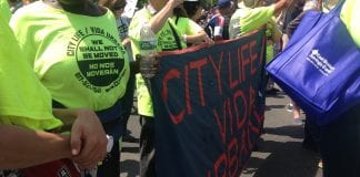 A dark-skinned woman in the foreground and two light-skinned men are wearing neon yellow T-shirts reading "City Life Vida Urbana, No Nos Moveran, We Shall Not Be Moved" and marching with a black banner with "City Life Vida Urbana" in red letters.