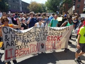 A crowd stands behind a multiracial group of people holding a cloth banner that reads "Capitalism, Racism, and Heteropatriarchy are making us sick. Join Liberation Health."