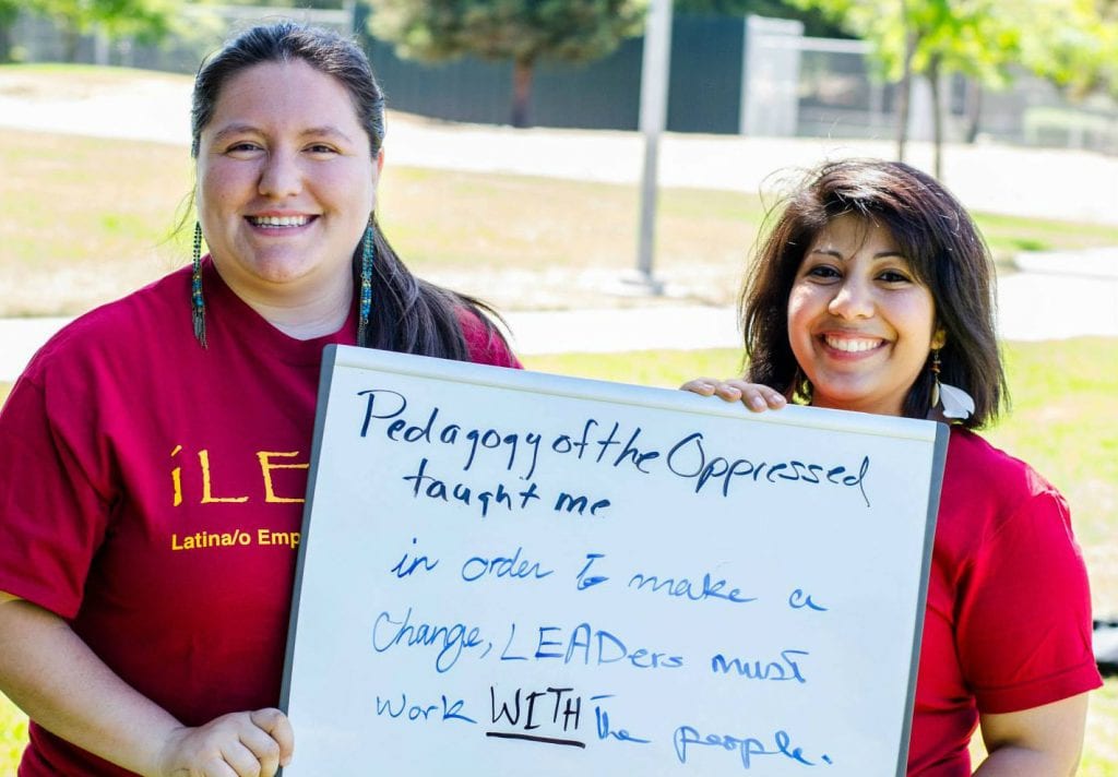 Two woman hold a sign that reads, "Pedagogy of the Oppressed Taught me ..."