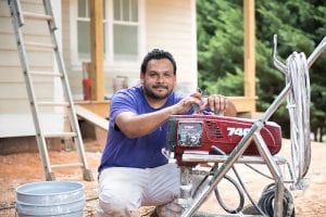 A man, owner of King of Kings Painting in North Carolina, received a business loan for his company. In this image, he kneels in front of a home he's painting along with his tools and paint.