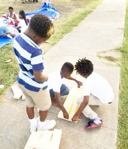 3 children measuring a board laid on a sidewalk.