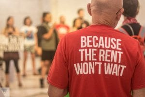 Close-up of the back of a T-shirt that says "Because the rent won't wait." Ways to help with the high cost of rent include rent control, rental assistance, and universal vouchers.