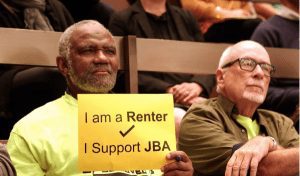 Man holding a yellow sign that reads "I am a Renter/I Support JBA"