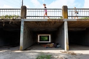 Two people sleeping beneath a pedestrian overpass in Bouldin Creek, Austin. A person walks above.