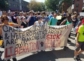 Members of Boston Liberation Health hold a banner that reads, "Capitalism, racism, and heteropatriachy are making us sick. Join Liberation Health."