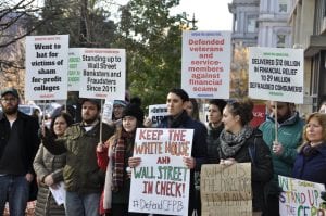 A group of men and women stand outside holding signs that defend the Consumer Financial Protection Bureau. A sign in the front center reads, "Keep the White House and Wall Street in check. #DefendCFBP"