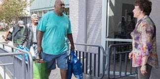 An African-American man in blue T-shirt and shorts, carry full shopping bags approaches the top of a ramp, where he is greeting by a white woman in a multicolored blouse. Two other people with belongings follow him up the ramp.