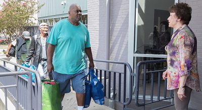 An African-American man in blue T-shirt and shorts, carry full shopping bags approaches the top of a ramp, where he is greeting by a white woman in a multicolored blouse. Two other people with belongings follow him up the ramp.