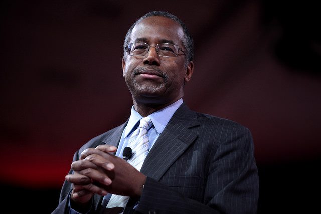 HUD Secretary Ben Carson smiles as his folds his hands in front of his chest.