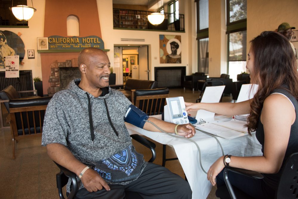 An African-American man gets his blood pressure checked at the California Hotel, an EBALDC development that offers affordable apartments with community and commercial spaces.