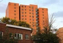 Multi-story public housing apartment building of reddish brick against a whitish-blue sky, looming over a darker, lower building in the foreground