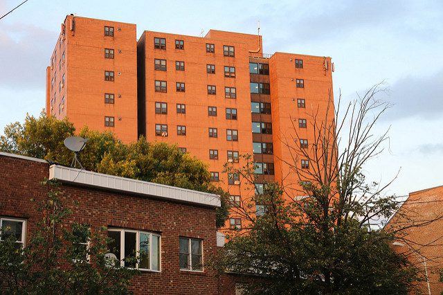 Multi-story public housing apartment building of reddish brick against a whitish-blue sky, looming over a darker, lower building in the foreground