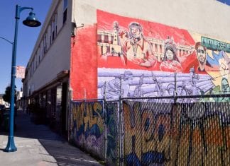 A building in East Oakland with colorful murals painted on the wall. A graffitied fence is to the right of the building.