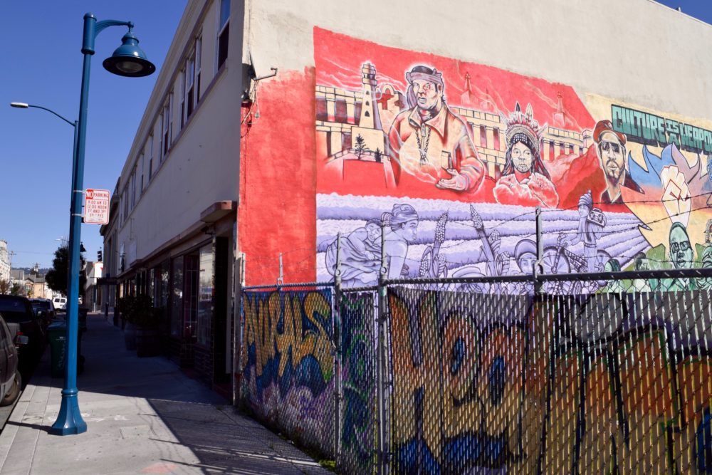 A building in East Oakland with colorful murals painted on the wall. A graffitied fence is to the right of the building.