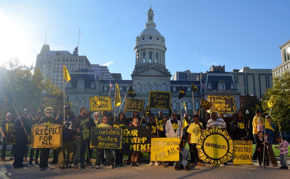 A group of people with yellow and black signs stand outside of Baltimore's City Hall. Some signs read "Respect for All," "Living Wages Now," and "Sueldos Justos Ahora."