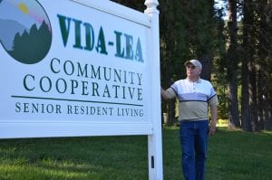 A white man stands near a sign that reads "Vida-Lea Community Cooperative Senior Resident Living."