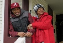 An African-American man and woman stand happily outside on their front porch. The woman is wearing a red coat and a black and white hate, while the man is reader a read Oakland A's hat and a black jacket.