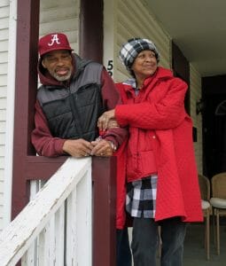 An African-American man and woman stand happily outside on their front porch. The woman is wearing a red coat and a black and white hate, while the man is reader a read Oakland A's hat and a black jacket.