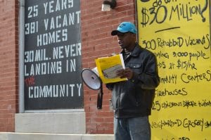 An African-American man speak through a bullhorn in front of a building with wording on it. The wording says "25 years, 381 vacant homes, 30 million never landing in the community."