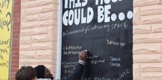A man and a woman stand in front of a chalkboard sign that reads "This House Could Be ..."The man is writing on the board, as many others have done. Some of the suggestions for what the house could be include a community gathering space and a senior center.