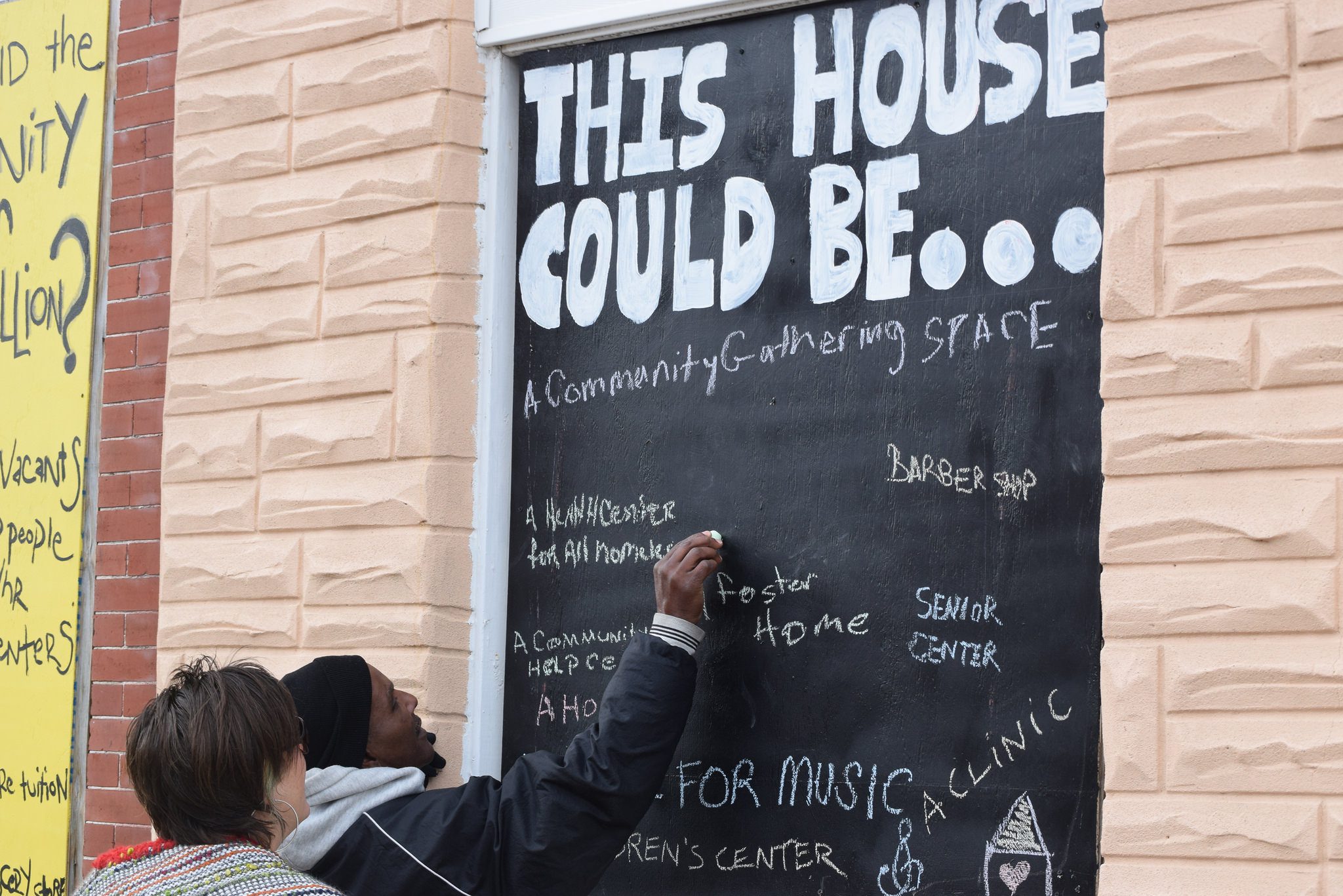 A man and a woman stand in front of a chalkboard sign that reads "This House Could Be ..."The man is writing on the board, as many others have done. Some of the suggestions for what the house could be include a community gathering space and a senior center.
