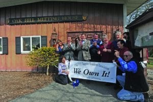 A group of residents stand outside of the Oak Hill Meeting House holding a sign that reads "We Own It." They are clapping and cheering.