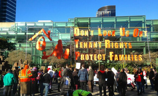 A crowd of people, backs to camera, stand befoer a four- or five-story building, holding tenants rights protest signs.