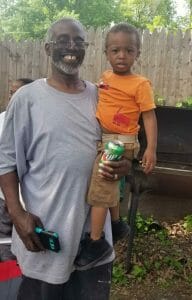 An African-American man in a gray shirt holds his young grandson in his arms. Brown faces eviction from his home due to its unsafe living conditions.