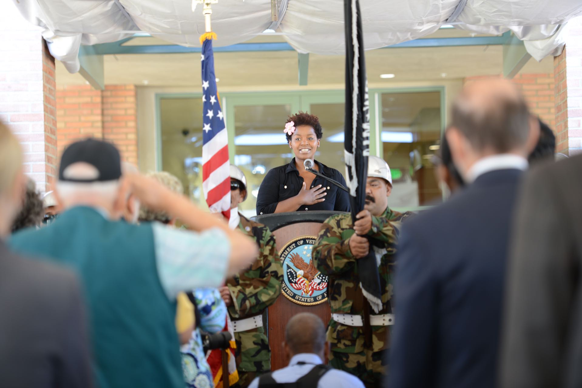 An African-American woman with a pink flower in her hair stands at a podium with a Veteran's Affairs seal on it with her hand on her heart, facing a crowd. An American flag is to her side.