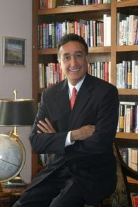 semiformal portrait of Henry Cisneros, seated on the side of his desk, with a wall of bookshelves behind him