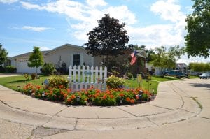 A small white fense with a sign that reads "welcome" and red flowers frame a resident-owned community in Wisconsin.