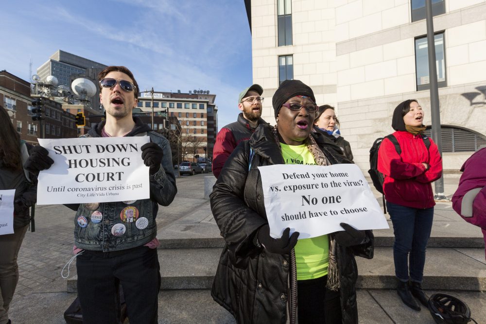 A group of people stand three feet apart, mouths open as if shouting, outside a building that looks like a court. In the foreground a young white man in sunglasses with political buttons on his jacket holds a sign reading "Shut down housing court until coronavirus is past." In the center, a Black woman with a neon yellow T-shirt and black jacket holds a sign reading "Defend your home versus exposure to the virus? No one should have to choose."