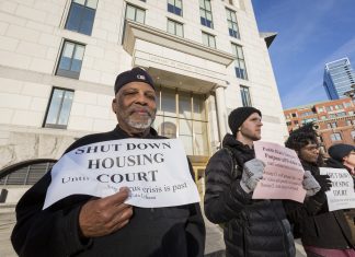 A group of people stand in a line outside a building a court building. In the foreground Black man with a white beard in a black jacket and cap holds a sign a sign reading "Shut down housing court until coronavirus is past."