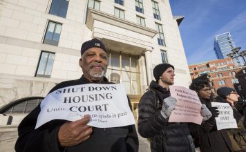 A group of people stand in a line outside a building a court building. In the foreground Black man with a white beard in a black jacket and cap holds a sign a sign reading "Shut down housing court until coronavirus is past."