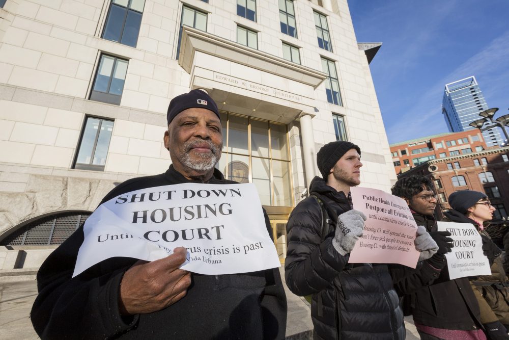 A group of people stand in a line outside a building a court building. In the foreground Black man with a white beard in a black jacket and cap holds a sign a sign reading "Shut down housing court until coronavirus is past."